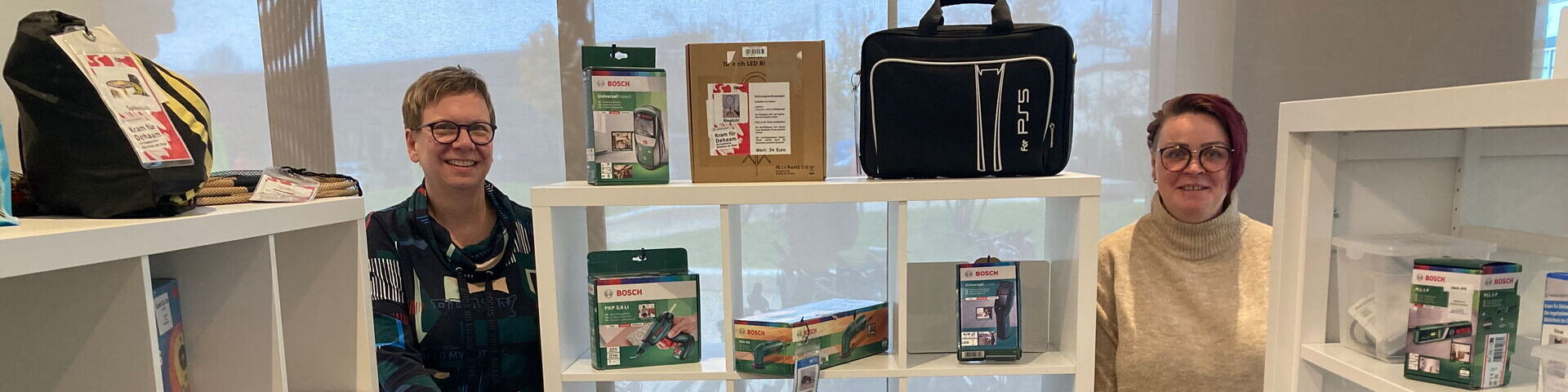 Das Bild zeigt zwei Personen, die vor Regalen mit verschiedenen Produkten stehen. Die Regale enthalten Artikel wie Taschen, Kisten und andere verpackte Waren.