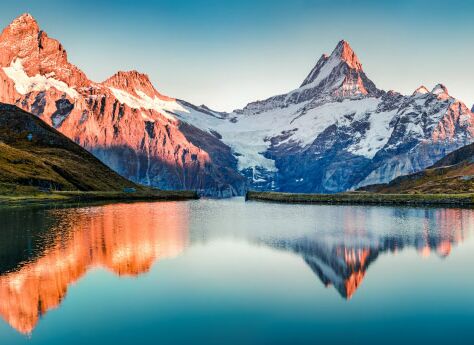 Das Bild zeigt eine ruhige Berglandschaft mit einem stillen See im Vordergrund. Der See spiegelt die umliegenden schneebedeckten Berge und den klaren Himmel wider. Die Berge sind vom warmen Licht des Sonnenaufgangs beleuchtet.
