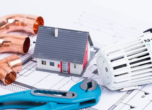 Auf dem Bild ist ein Bauplan zu sehen. Darauf ist ein kleines Häuschen, eine Zange, Rohre und ein Heizungsthermostat platziert.