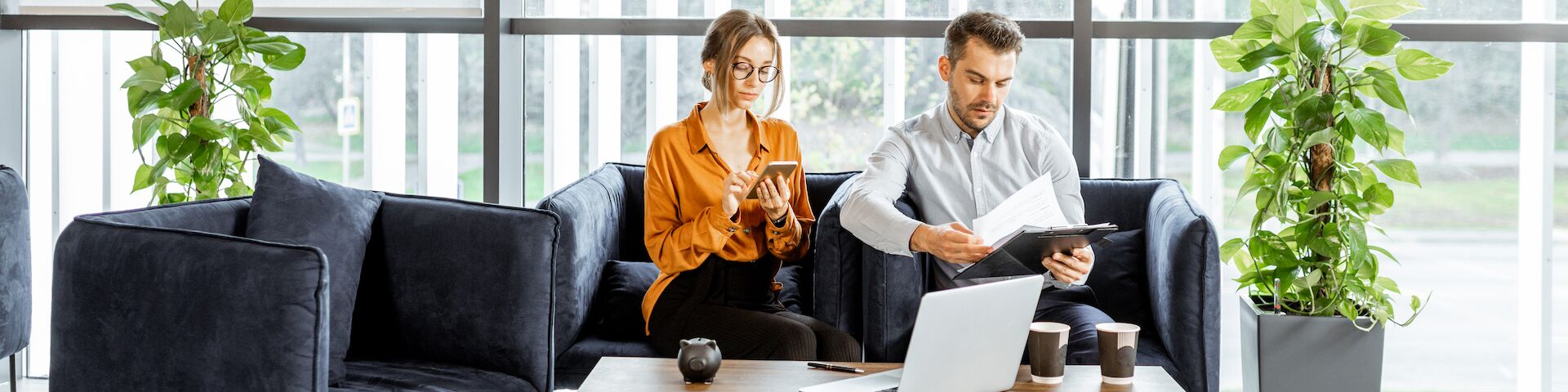 Eine Frau und ein Mann sitzen auf einem dunkelblauen Sofa in einem Büro. Die Frau trägt ein orangefarbenes Hemd und tippt auf einem Smartphone, während der Mann einige Papiere in der Hand hält.