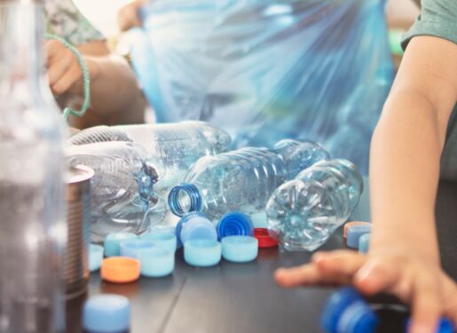 Das Bild zeigt eine Gruppe von Menschen, die verschiedene Plastikflaschen und -deckel auf einem Tisch sortieren. Auf dem Tisch liegen mehrere Plastikflaschen, Flaschendeckel und andere recycelbare Materialien verstreut.