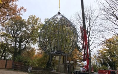 Das Bild zeigt eine große, kugelförmige Metallstruktur, die von einem Kran angehoben wird. Die Struktur scheint aus miteinander verbundenen Metallstangen zu bestehen, die ein geometrisches Muster bilden. Die Spitze der Ingelummer Kerz.