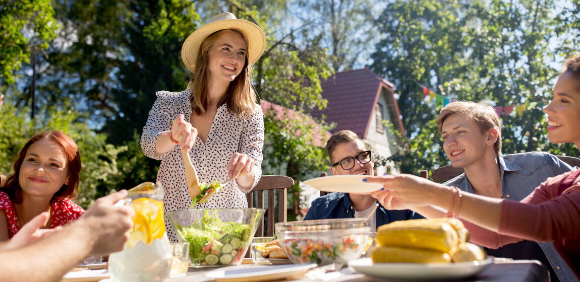 Zu sehen ist eine Familie draußen im Garten beim Sommergrillen. Es gibt Salate und Maiskolben. Im Hintergrund befindet sich ein Haus und Bäume.