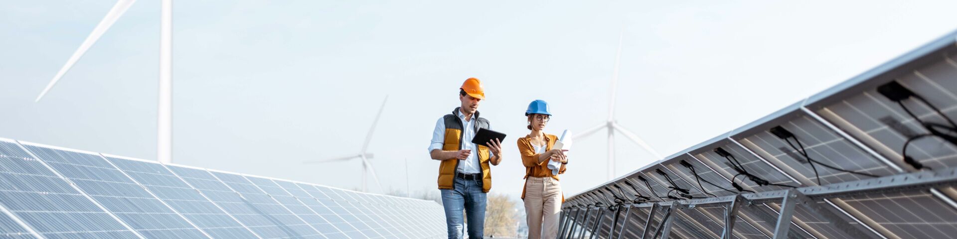 Das Bild zeigt zwei Personen, die Sicherheitshelme und Westen tragen und zwischen Reihen von Solarpaneelen auf einer Solarfarm gehen. Im Hintergrund sind Windturbinen sichtbar.