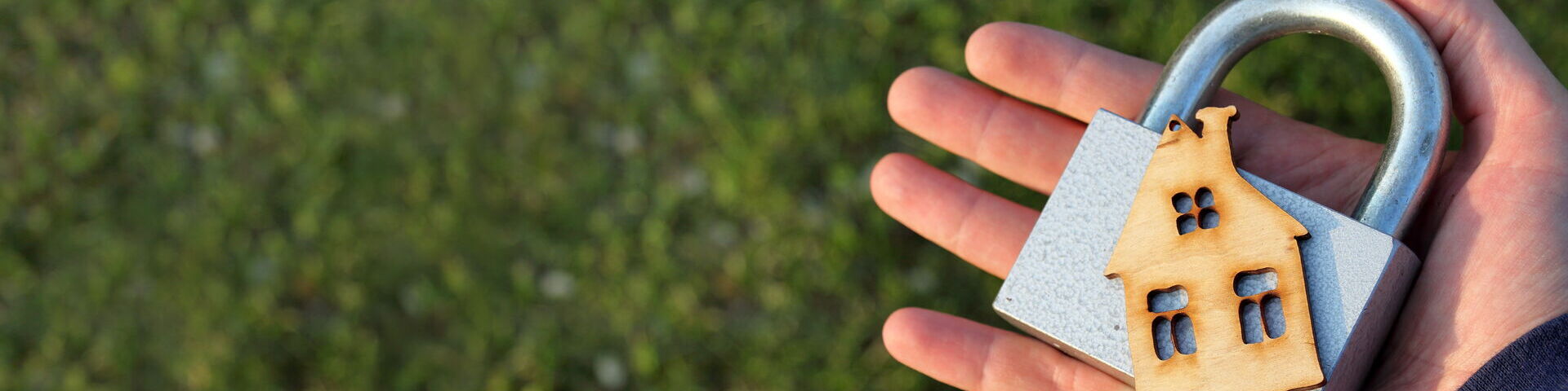 Das Bild zeigt eine Hand, die ein großes Vorhängeschloss hält, an dem ein kleines Holzhaus befestigt ist. Der Hintergrund besteht aus grünem Gras.