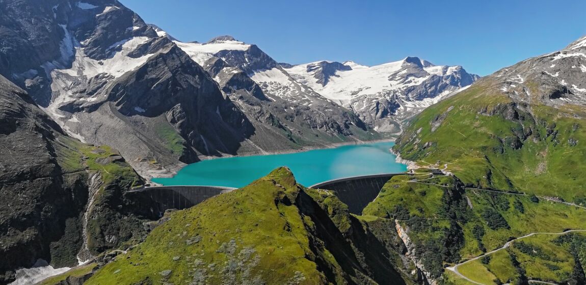 Das Bild zeigt eine malerische Landschaft mit schneebedeckten Berggipfeln im Hintergrund. In der Mitte des Bildes befindet sich ein türkisfarbener See, der von steilen Bergen umgeben ist. Der See scheint von einem Damm gehalten zu werden.