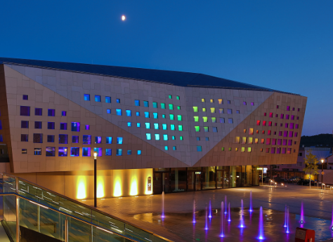 Die kING - Kultur- und Kongresshalle Ingelheim in der Dämmerung mit einem Mond am Himmel mit regenbogenfarbigen kleinen Fenstern. Vor dem Gebäude befindet sich ein offener Platzbereich mit beleuchteten Wasserfontänen.