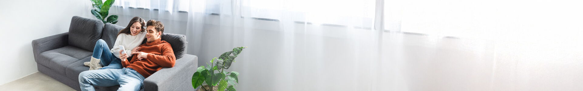 Im Bild zu sehen ist ein junges Pärchen. Sie sitzen auf einer grauen Couch. Gemeinsam schauen Sie auf ein Handy.