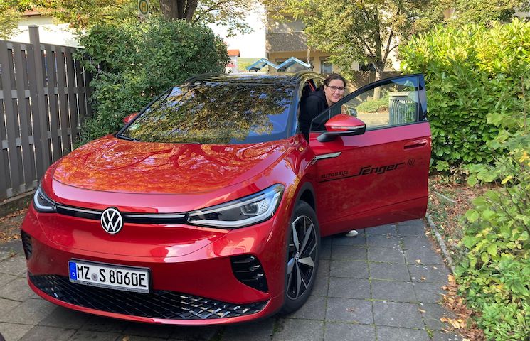 Das Bild zeigt ein rotes Elektrofahrzeug der Marke Volkswagen, das in einer Einfahrt geparkt ist. Die Fahrertür ist geöffnet, und eine Person sitzt im Auto.