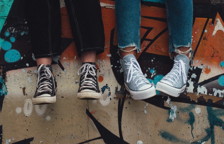 Das Bild zeigt zwei Paar Beine, die Sneakers tragen und auf einer mit Graffiti bedeckten Mauer sitzen. Das linke Paar Beine trägt schwarze Sneakers. Das rechte Paar Beine trägt hellgraue Sneakers.