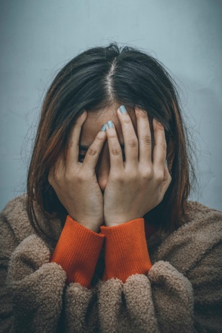 Das Bild zeigt eine Person mit langen Haaren, die ihr Gesicht mit beiden Händen bedeckt. Die Person trägt ein orangefarbenes Langarmshirt und eine braune, flauschige Jacke. Der Hintergrund ist schlicht und grau.