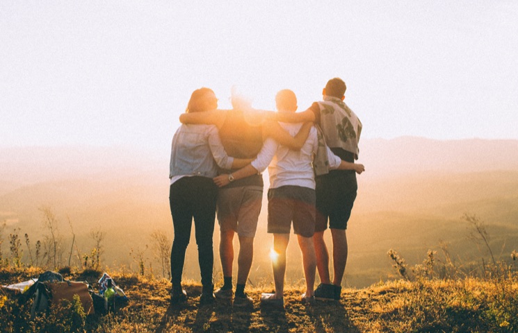 Das Bild zeigt vier Personen, die auf einem Hügel während des Sonnenuntergangs stehen. Sie haben ihre Arme umeinander gelegt, schauen von der Kamera weg und in Richtung Horizont. Die Sonne steht tief am Himmel und wirft ein warmes, goldenes Licht.