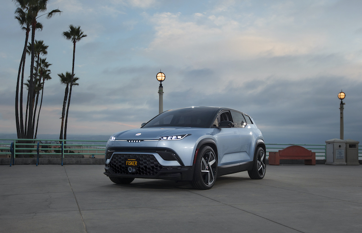 Das Bild zeigt einen hellblauen elektrischen SUV - Fisker Ocean One, der auf einer Betonfläche in der Nähe eines Küstengebiets geparkt ist. Das Fahrzeug hat moderne, schlanke Linien und markante LED-Scheinwerfer. Im Hintergrund gibt es hohe Palmen.
