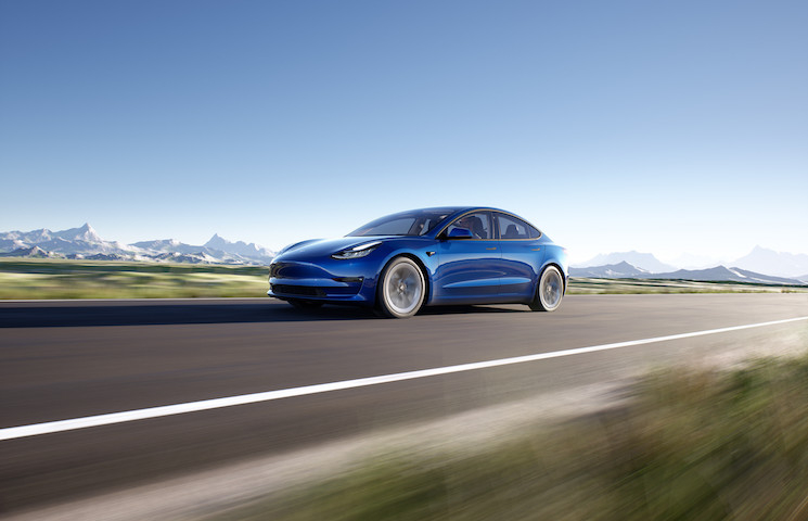 Das Bild zeigt ein blaues Elektroauto, den Tesla Model 3, das auf einer Straße fährt, mit Bergen im Hintergrund. Das Auto hat ein schlankes und modernes Design, mit einer glatten Karosserie und aerodynamischen Merkmalen.