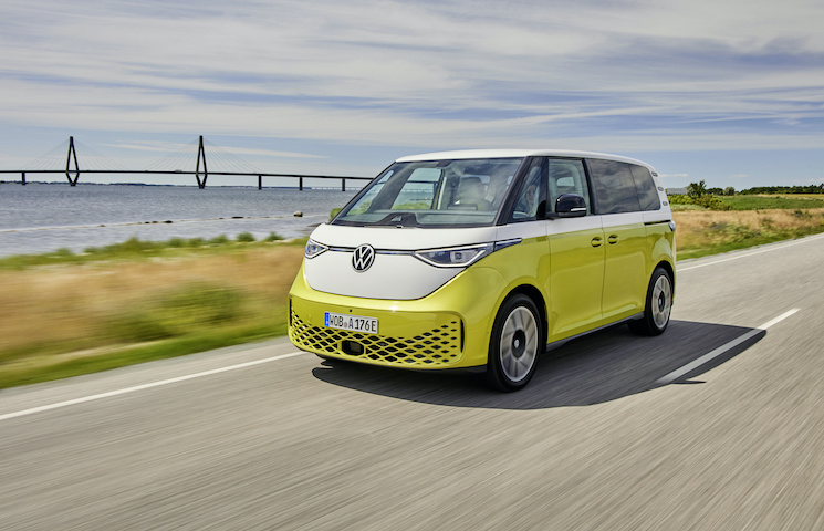 Das Bild zeigt einen gelb-weißen Volkswagen-Bus, den VW-ID-Buzz, der auf einer Straße in der Nähe eines Gewässers fährt. Im Hintergrund ist eine Brücke zu sehen.