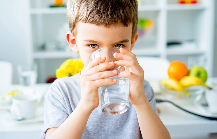 Ein Junge mit kurzen brauen Haaren hält mit beiden Händen ein Wasserglas und trinkt.