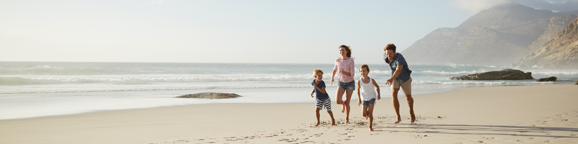 Eine Familie mit vier Personen, die am Strand entlanglaufen. Im Hintergrund sieht man das Meer mit sanften Wellen und entfernten Bergen unter einem klaren Himmel.