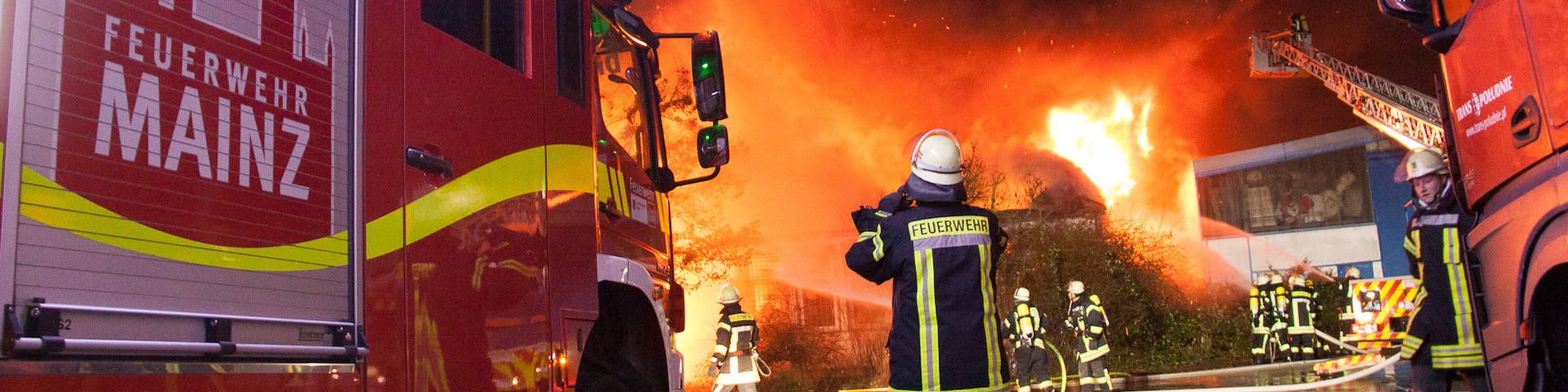 Eine Gruppe von Feuerwehrleuten, die aktiv daran arbeiten, ein großes Feuer zu löschen. Es sind zwei Feuerwehrautos mit der Aufschrift "FEUERWEHR MAINZ" zu sehen. Die Feuerwehrleute tragen Schutzkleidung, einschließlich Helmen und reflektierenden Jacken.