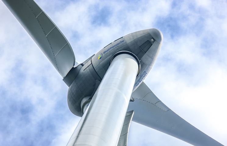 Nahaufnahme einer Windturbine vor einem blauen Himmel mit einigen Wolken. Der Fokus liegt auf der Gondel (das Gehäuse, das den Generator und andere Komponenten enthält) und dem Ansatz der Rotorblätter.