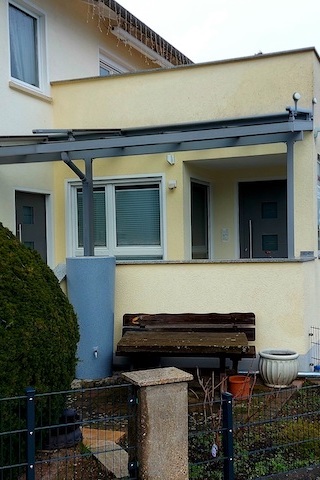 Vordereingang eines Hauses. Das Haus hat einen kleinen Verandabereich mit einer Holzbank und einigen Topfpflanzen. Über der Veranda befindet sich ein Vordach, das von Metallträgern gestützt wird.