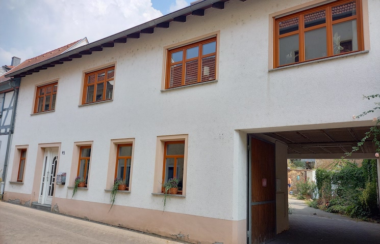 Zweistöckiges weißes Gebäude mit braun gerahmten Fenstern. Im Erdgeschoss befinden sich 4 Fenster, die jeweils mit Blumen versehen sind. Die Eingangstür befindet sich auf der links und auf der rechten Seite führt ein Durchgang nach hinten.