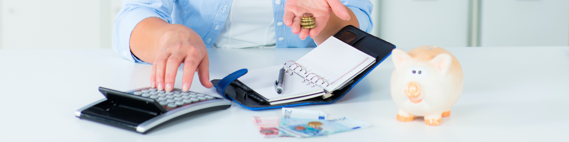Eine Person, die mit einer Hand einen Taschenrechner benutzt, während sie in der anderen Hand Münzen hält. Auf dem Tisch liegen ein offenes Notizbuch mit einem Stift, ein Sparschwein und einige Geldscheine.