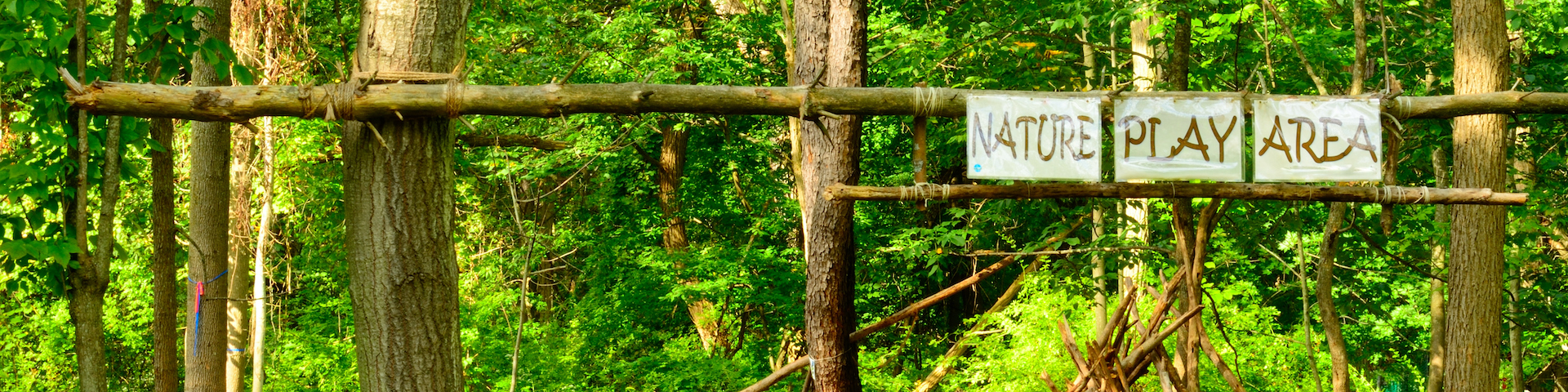 Eine Holzstruktur in einem bewaldeten Gebiet mit einem Schild, auf dem "NATURE PLAY AREA" steht. Die Struktur besteht aus Baumstämmen und Ästen, und der Hintergrund besteht aus dichtem grünen Laub.