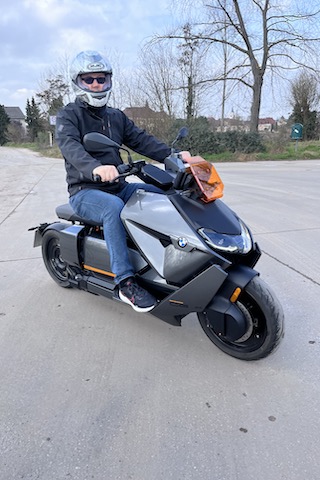 Frank Hilbrath auf einem modernen, schwarz-grauen Elektroroller mit einer orangefarbenen Windschutzscheibe. Die Person trägt einen Helm, eine dunkle Jacke, blaue Jeans und schwarze Schuhe. Im Hintergrund sind Bäume und Häuser in der Ferne zu sehen.