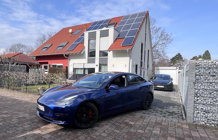 Ein modernes Haus mit Solarpaneelen auf dem Dach. Vor dem Haus steht ein blaues Elektroauto in der Einfahrt. Im Hintergrund ist ein weiteres schwarzes Auto zu sehen, das ebenfalls in der Einfahrt geparkt ist.