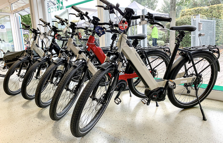 Eine Reihe von Elektrofahrrädern (E-Bikes), die drinnen ausgestellt sind. Die Fahrräder haben dicke Reifen, Vorderradaufhängungen und sichtbare Batteriepacks, die in die Rahmen integriert sind.