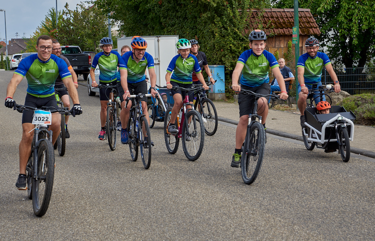 Eine Gruppe von Radfahrern, die auf einer Straße fahren. Insgesamt sind acht Radfahrer zu sehen, alle tragen passende blau-grüne Trikots. Ein Radfahrer zieht einen Anhänger mit einer Abdeckung.