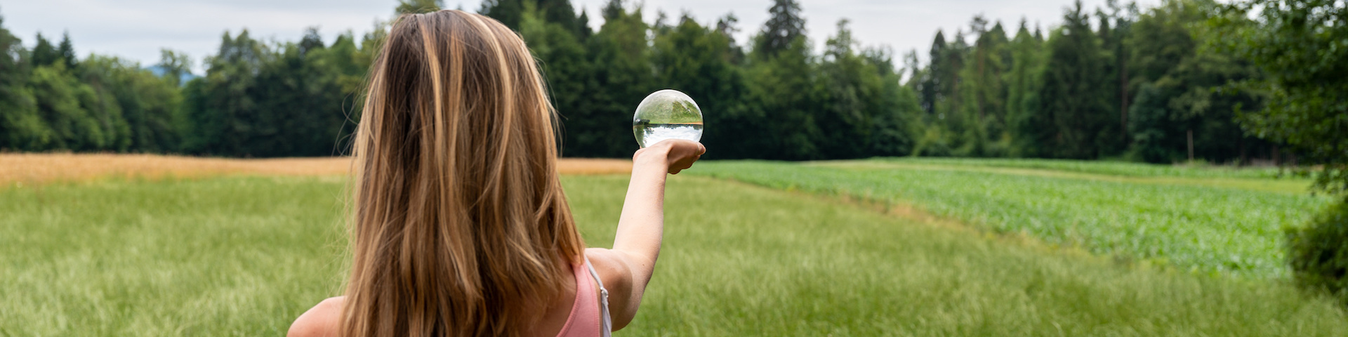 Eine Frau mit langen Haaren steht in einem grasbewachsenen Feld und hält ein klares, kugelförmiges Objekt auf Armlänge. Im Hintergrund sind Bäume und ein offener Himmel zu sehen.