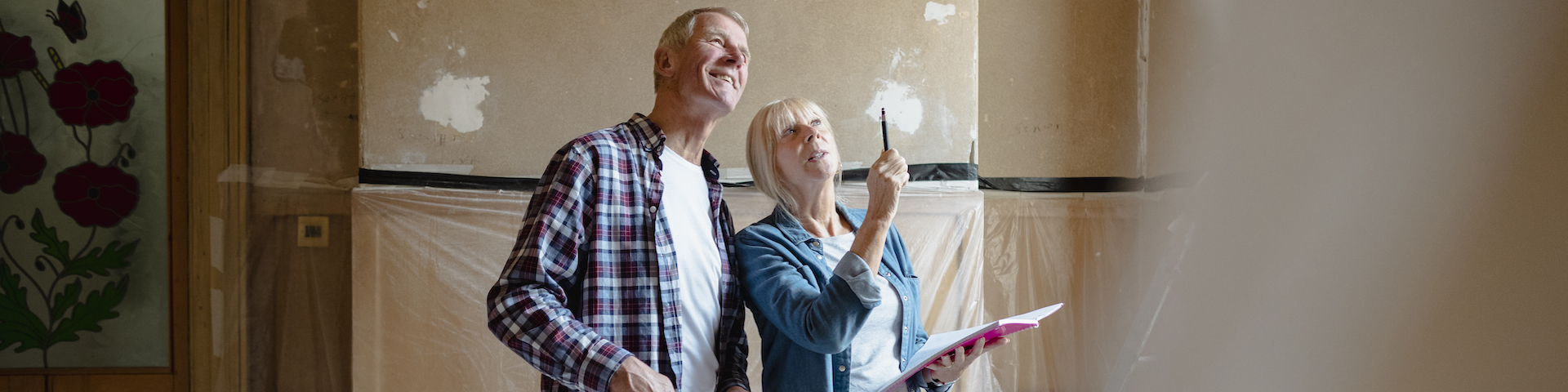 Eine Frau und ein Mann stehen in einem Raum, der renoviert wird. Die Frau hält ein Notizbuch und zeigt auf etwas an der Wand, während der Mann in die gleiche Richtung schaut. Die Wände haben Putzstellen.