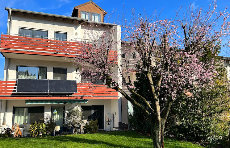 Das Bild zeigt ein mehrstöckiges Wohngebäude mit Balkonen. Die Wände des Gebäudes sind weiß und die Geländer der Balkone sind rot. Auf einem der Balkone sind Solarpaneele installiert. Vor dem Gebäude befindet sich ein gepflegter Rasen.