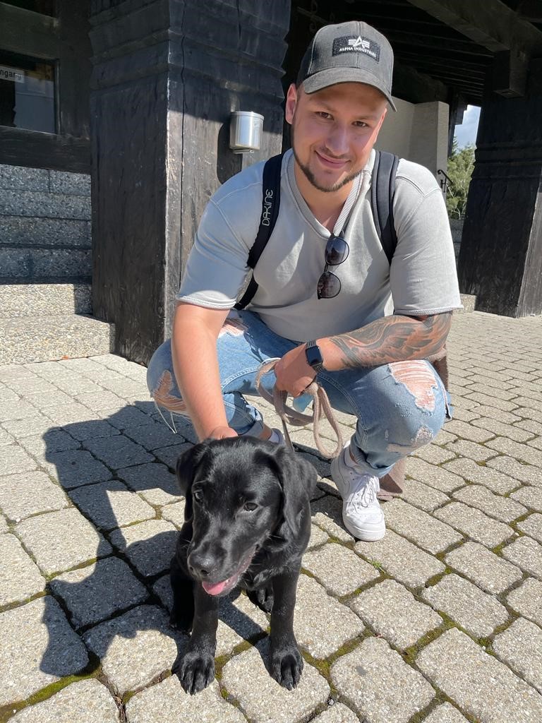 Paul Saraev, der auf einer gepflasterten Fläche hockt und die Leine seines schwarzen Hundes hält. Die Person trägt ein graues Shirt, zerrissene Jeans, weiße Sneakers und eine schwarze Kappe.