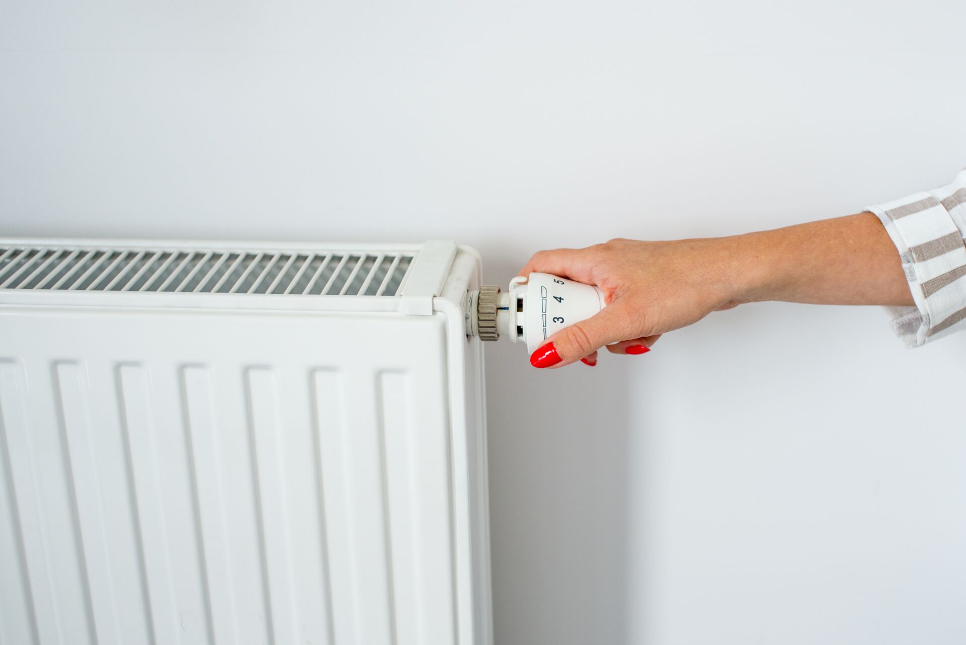 Das Bild zeigt eine Hand, die den Thermostat an einem weißen Heizkörper einstellt. Die Hand hat rot lackierte Fingernägel und dreht den Knopf an der Seite des Heizkörpers, der Zahlen und Symbole zur Anzeige der verschiedenen Temperatureinstellungen hat.