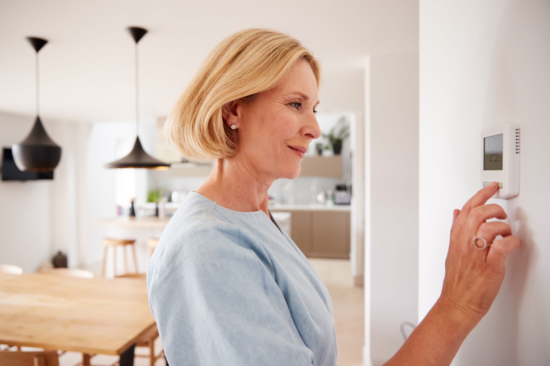 Eine Frau mit kurzen blonden Haaren, die ein hellblaues Hemd trägt und einen Thermostat an der Wand einstellt. Im Hintergrund sieht man eine moderne Küche und einen Essbereich mit Holzmöbeln und schwarzen Pendelleuchten.