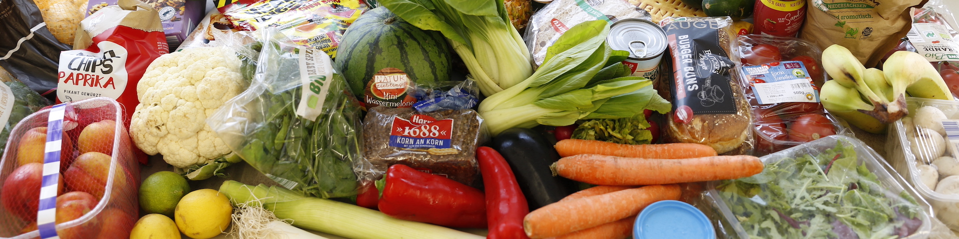 Das Bild zeigt eine Vielzahl frischer Gemüse, Früchte und anderer Lebensmittel, die auf einer Fläche ausgebreitet sind. Sichtbare Artikel umfassen Blumenkohl, Karotten, Pak Choi, rote Paprika, Bananen, Äpfel, Zitronen, Limetten und Blattgemüse.
