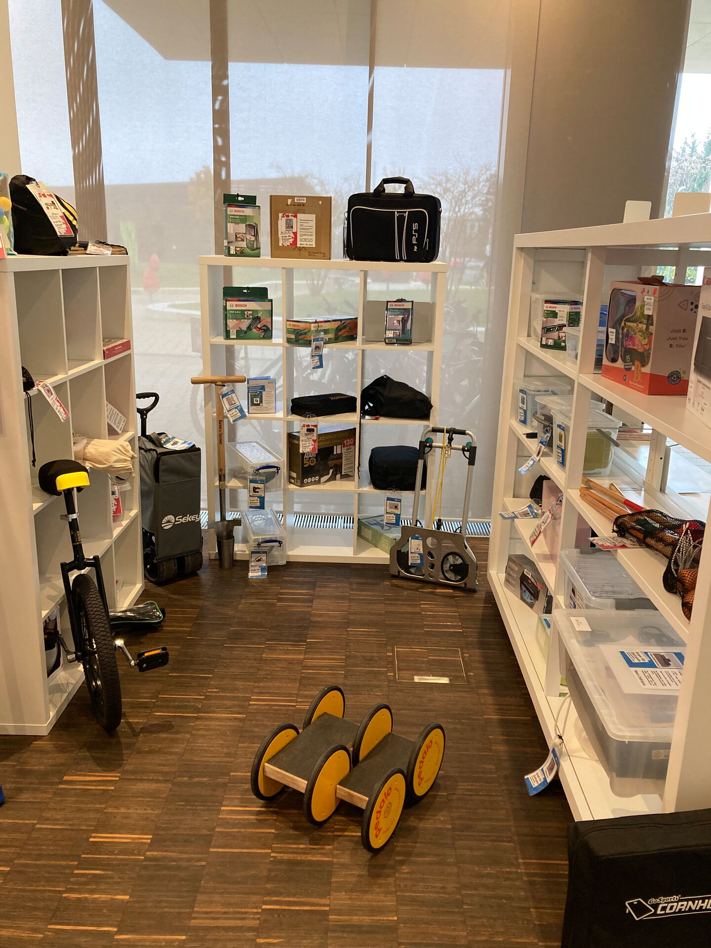 Mediathek der Dinge, das Bild zeigt einen Raum mit mehreren weißen Regalen, die mit verschiedenen Gegenständen gefüllt sind. Die Regale enthalten Kisten, Taschen, elektronische Geräte und andere diverse Objekte.