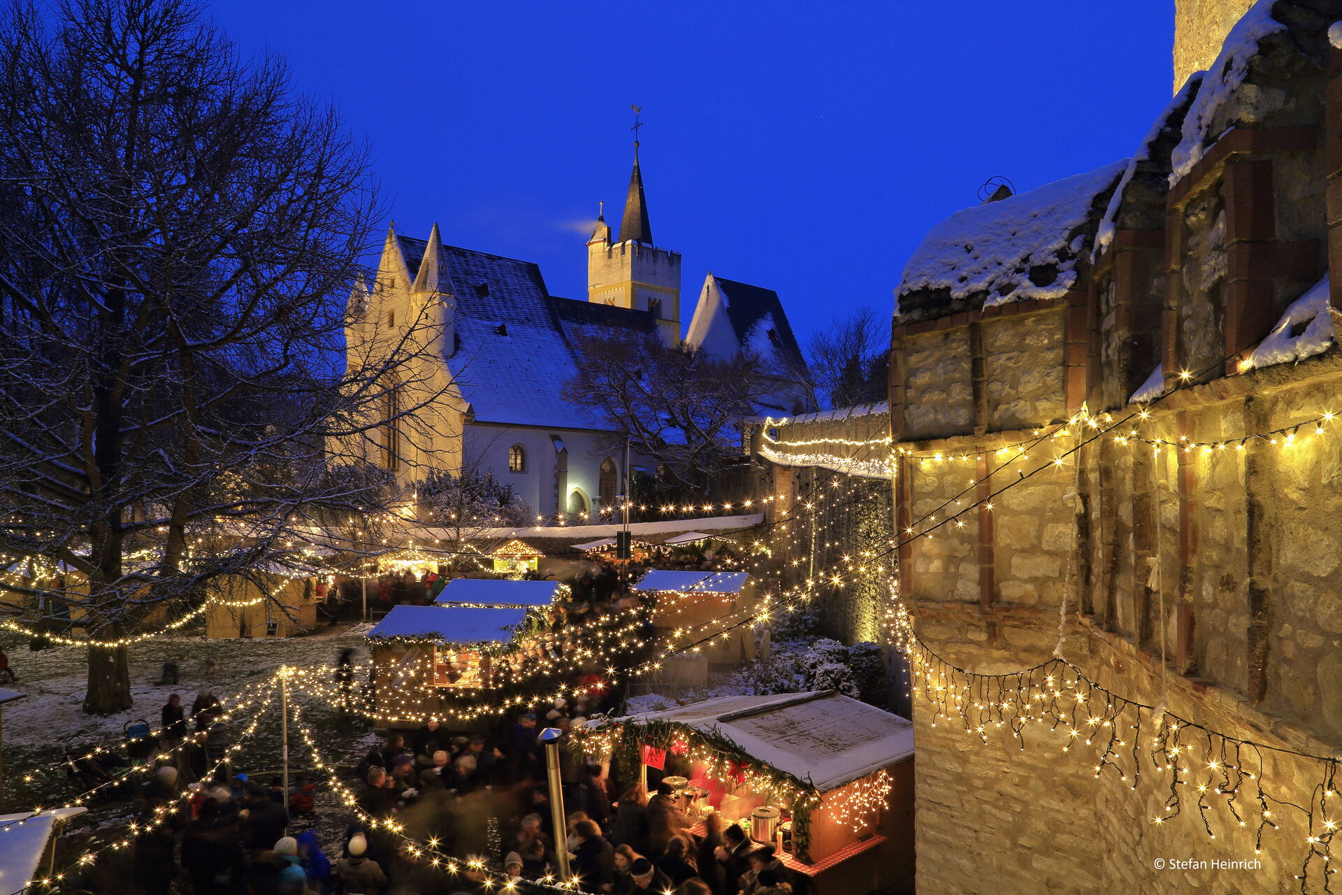 Das Bild zeigt eine festliche Weihnachtsmarktszene am Abend mit zahlreichen Lichterketten, die den Bereich beleuchten. Es gibt mehrere kleine Holzstände, die mit Lichtern und Schnee dekoriert sind, und eine Menschenmenge, die zwischen i