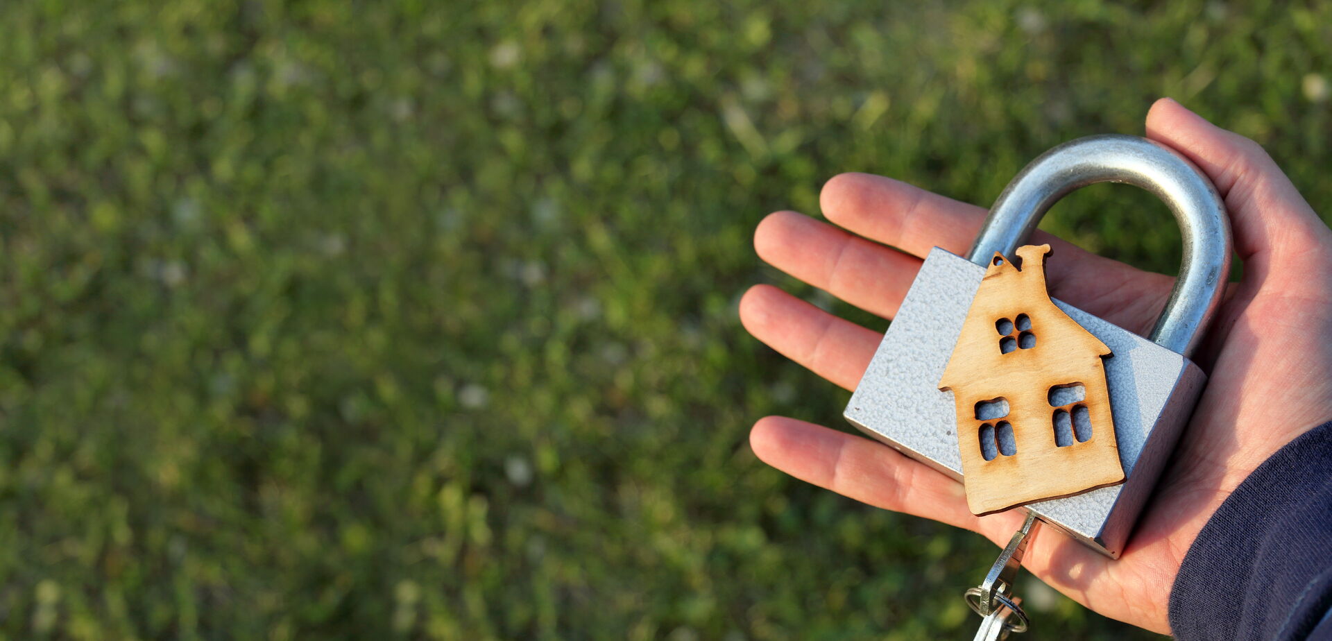 Das Bild zeigt eine Hand, die ein großes Vorhängeschloss hält, an dem ein kleines Holzhaus befestigt ist. Der Hintergrund besteht aus grünem Gras.