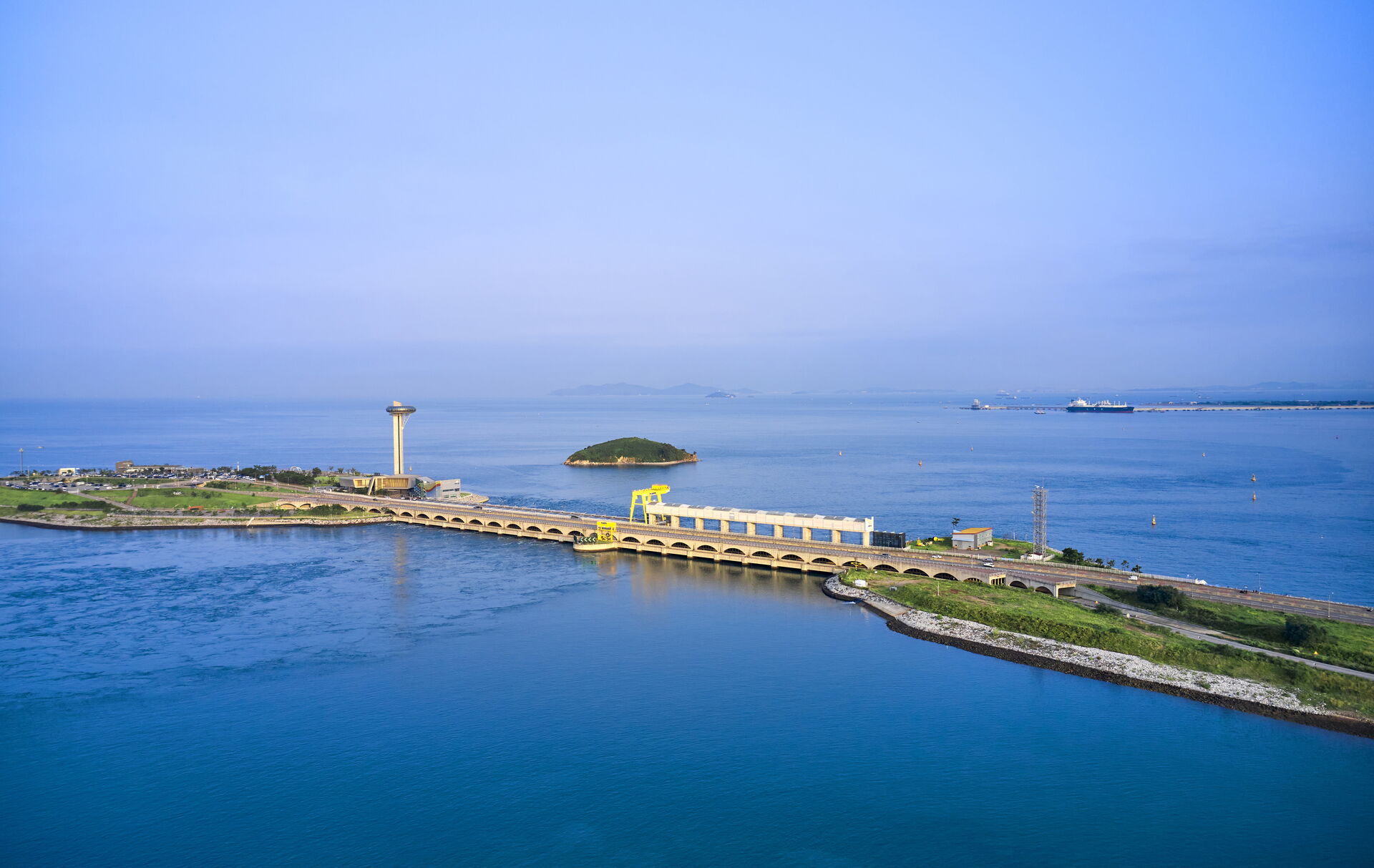 Das Bild zeigt eine Küstenregion mit einer langen Brücke, der sich über das Wasser erstreckt. Auf der linken Seite der Brücke befindet sich ein hoher Aussichtsturm, und in der Ferne ist eine kleine Insel zu sehen. Das Wasser ist ruhig.