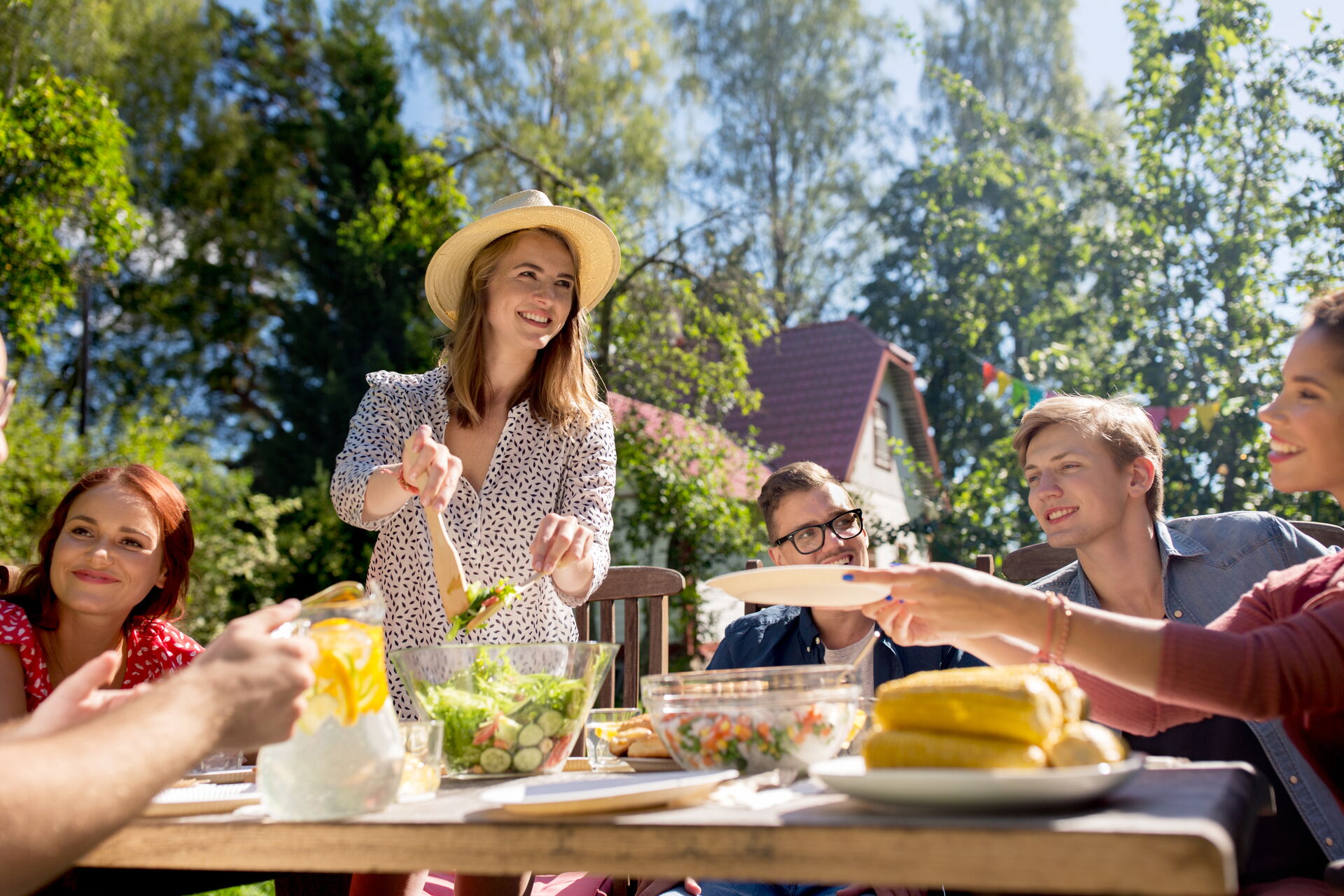 Zu sehen ist eine Familie draußen im Garten beim Sommergrillen. Es gibt Salate und Maiskolben. Im Hintergrund befindet sich ein Haus und Bäume.