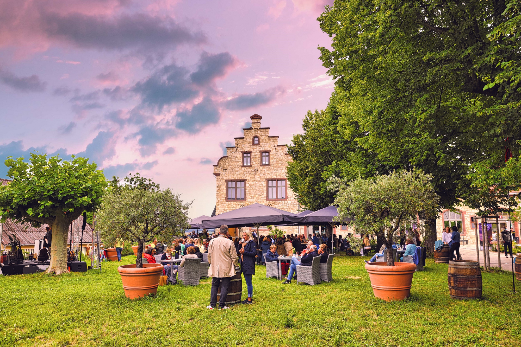 Zu sehen ist der Ingelheimer Winzerkeller. Bürger sitzen draußen im Garten und genießen die Stimmung bei einem Glas Wein.