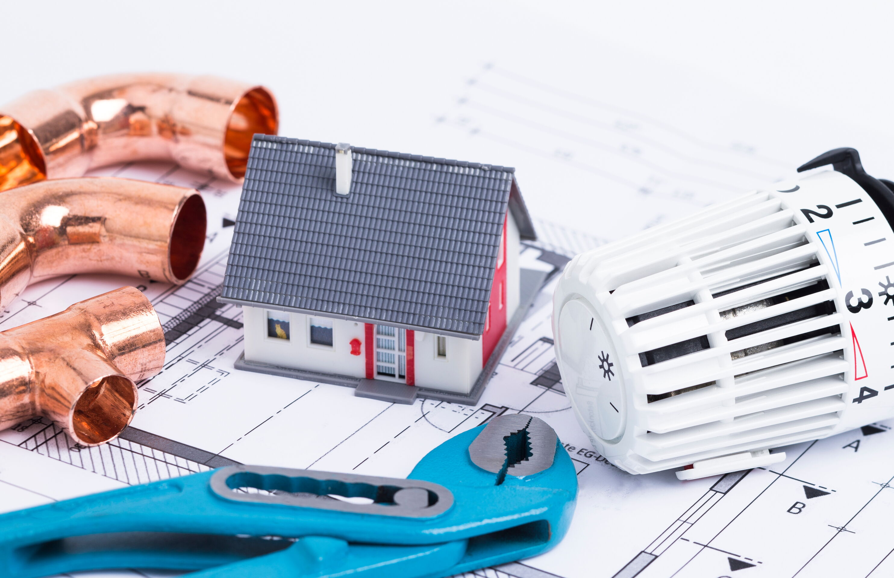 Auf dem Bild ist ein Bauplan zu sehen. Darauf ist ein kleines Häuschen, eine Zange, Rohre und ein Heizungsthermostat platziert.