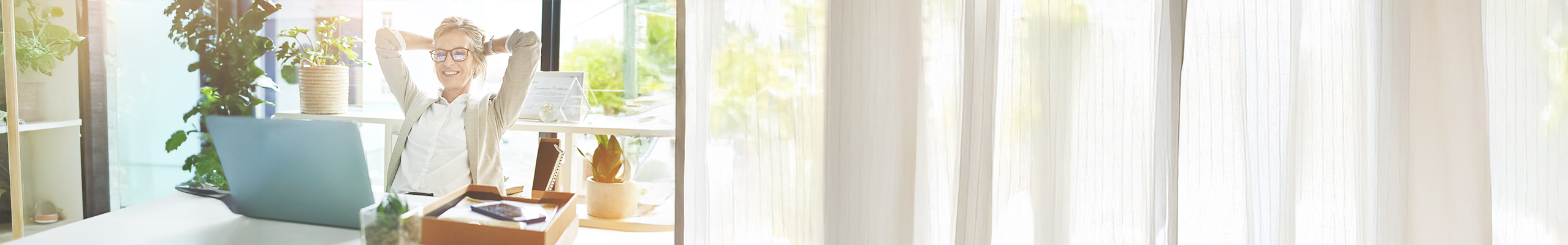 Zu sehen ist eine Frau mittleren Alters. Sie lächelt und lehnt sich entspannt in ihrem Schreibtischsessel zurück. Dabei blickt sie zufrieden auf ihren Laptop.