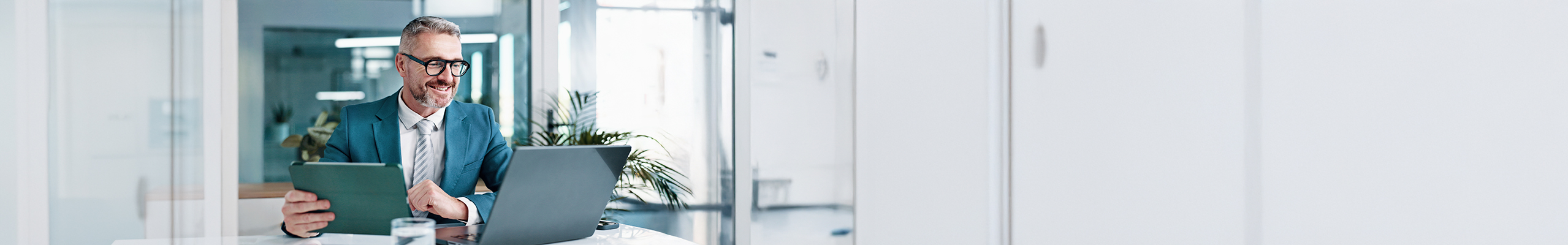 Ein Mann im Anzug sitzt in seinem Büro. Er trägt eine Brille und er schaut auf seinen Laptop. Vor ihm steht ein Glas Wasser.