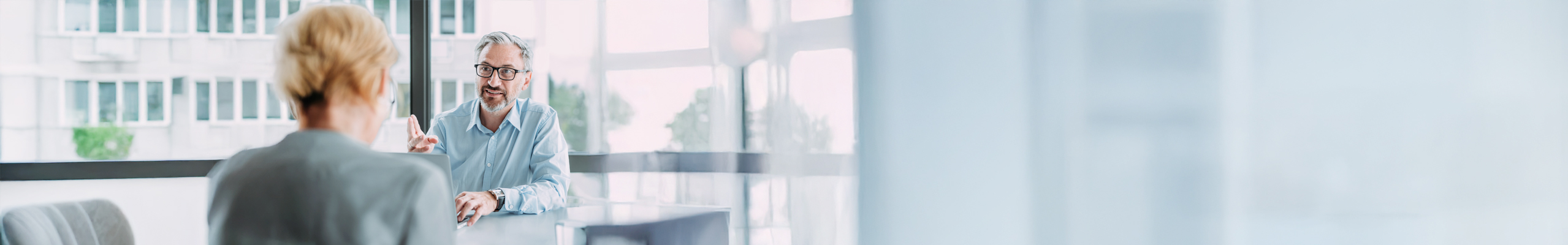 Zu sehen ist eine Beratungssituation in einem Bürokomplex. Der Berater trägt ein blaues Hemd und ist mittleren Alters. Die Kundin ist blond und nur von hinten zu sehen. Sie trägt eine graue Bluse, ihre blonden Haare trägt sie geschlossen.