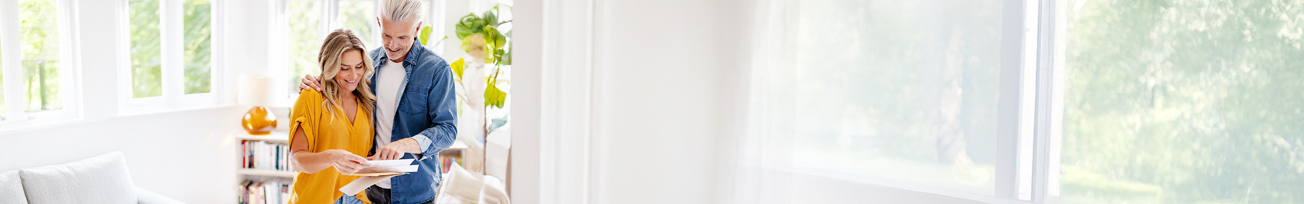 Ein Paar mittleren Alters steht in seinem Wohnzimmer. Sie trägt eine goldgelbe Bluse, er ein lockeres Jeanshemd. Die Einrichtung ist in weißen Tönen gehalten. Gemeinsam blicken sie zufrieden auf ein paar Dokumente.