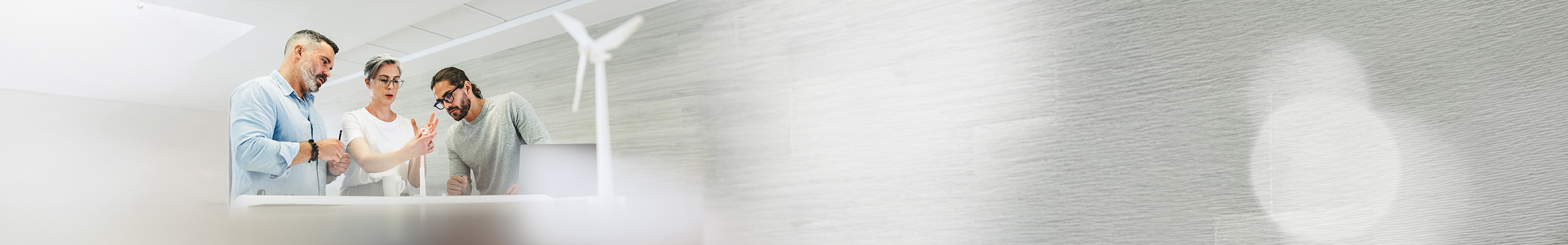 Drei Kollegen, davon zwei Männer und eine Frau, besprechen ein Projekt. Das komplette Porträt ist in Grau- und Weißtönen gehalten. Vor Ihnen steht kleines, weißes Windrad auf dem Tisch.